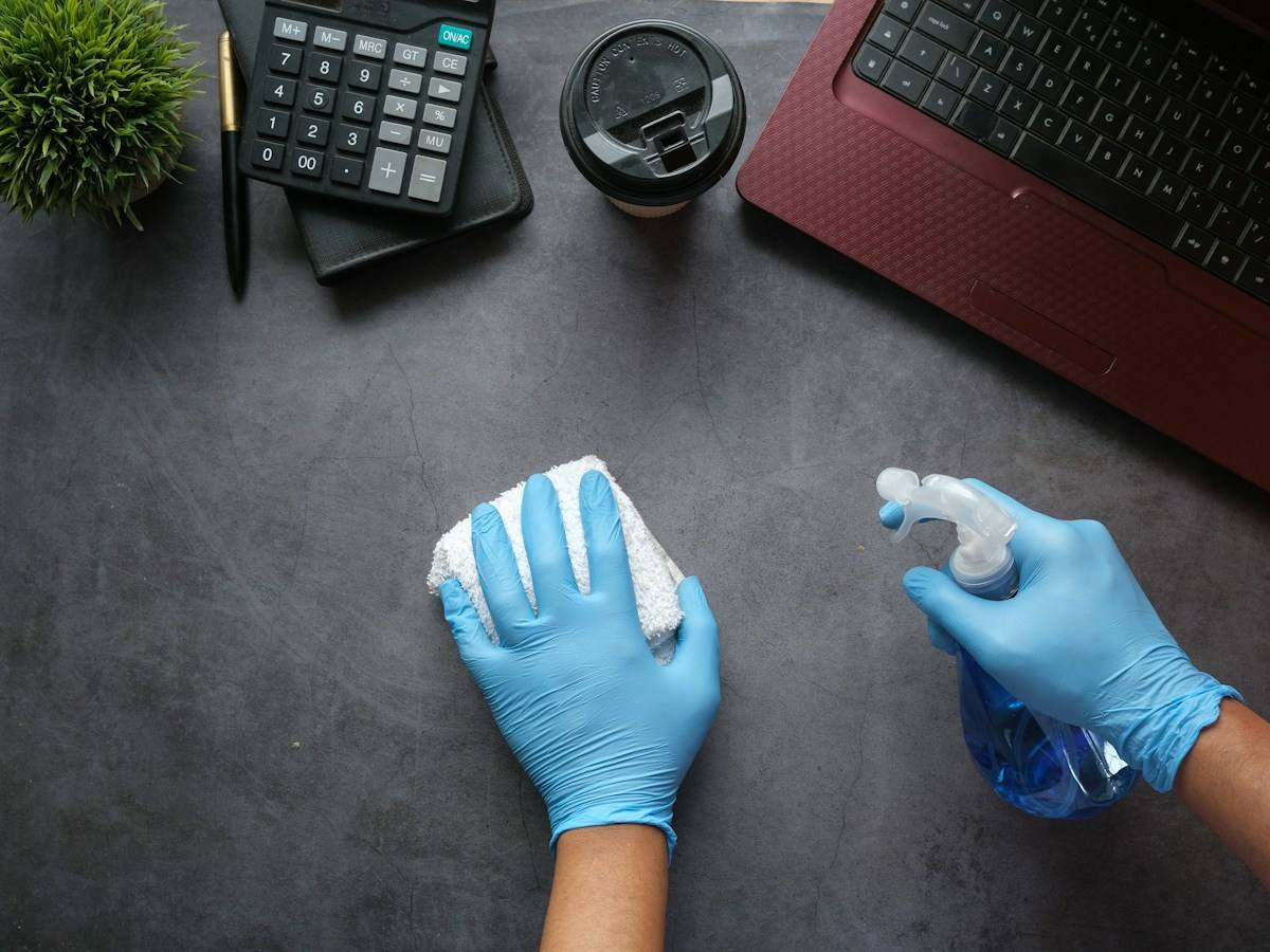 Mâini în mănuși de protecție care dezinfectează un birou cu spray și lavetă